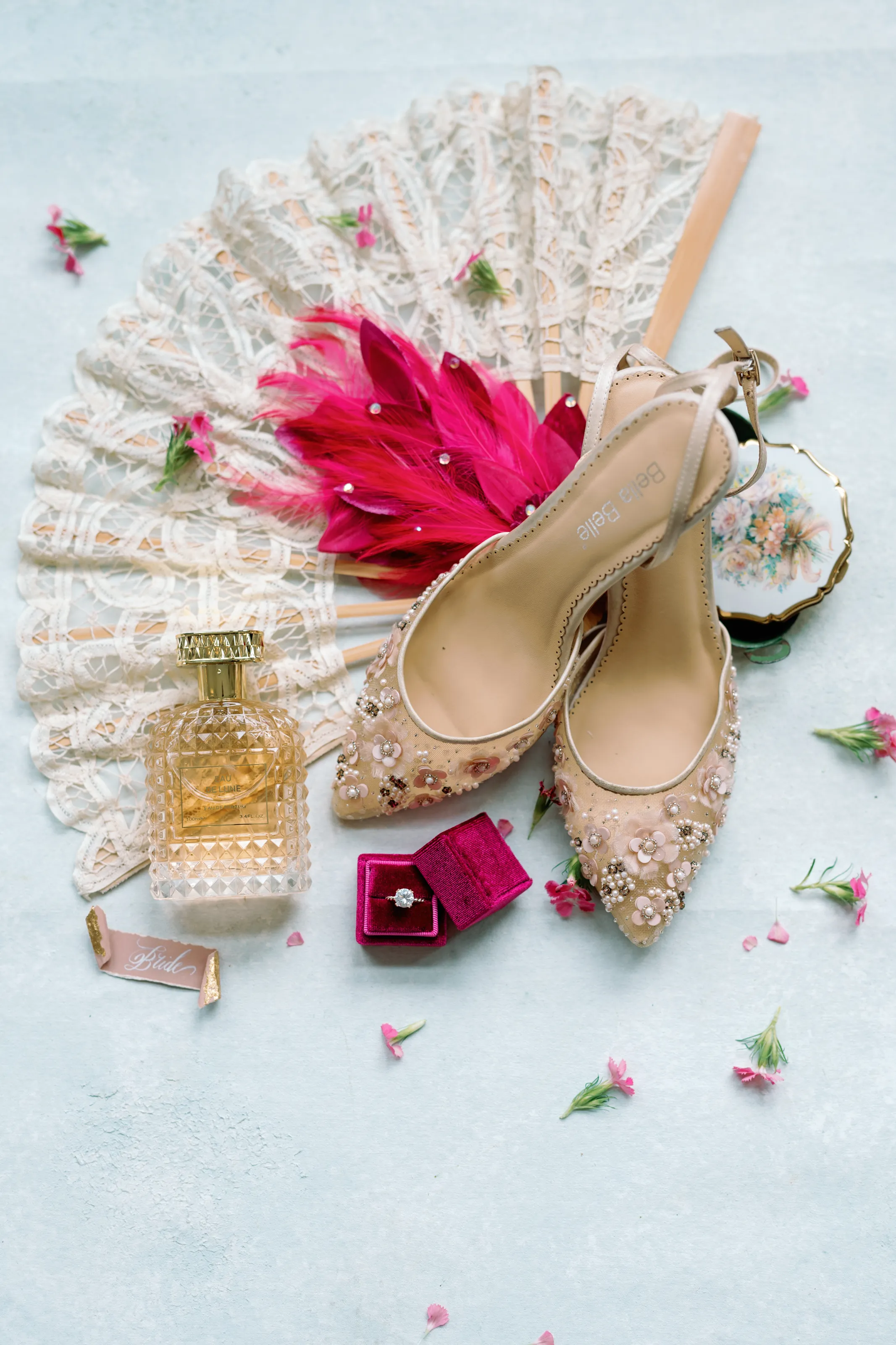 Bridal details flatlay: beaded heels, engagement ring, a lace fan, and perfume styled with loose pink petals