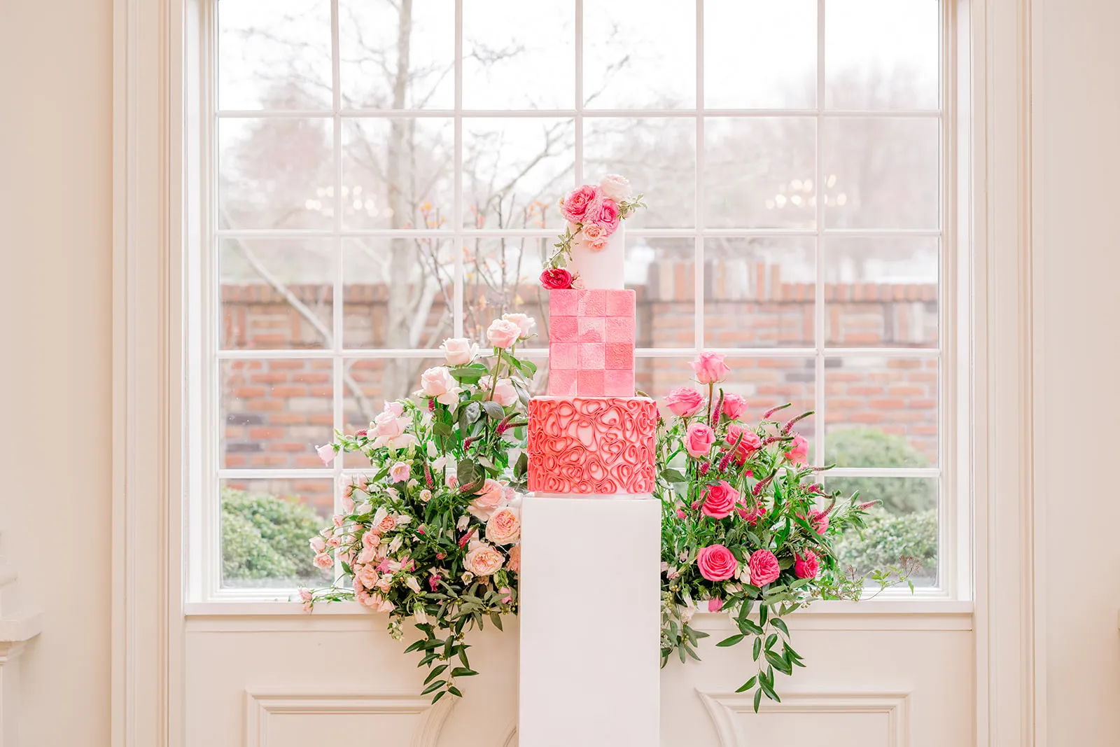 An ombré pink tiered wedding cake accented with garden roses, framed by a window and floral urns