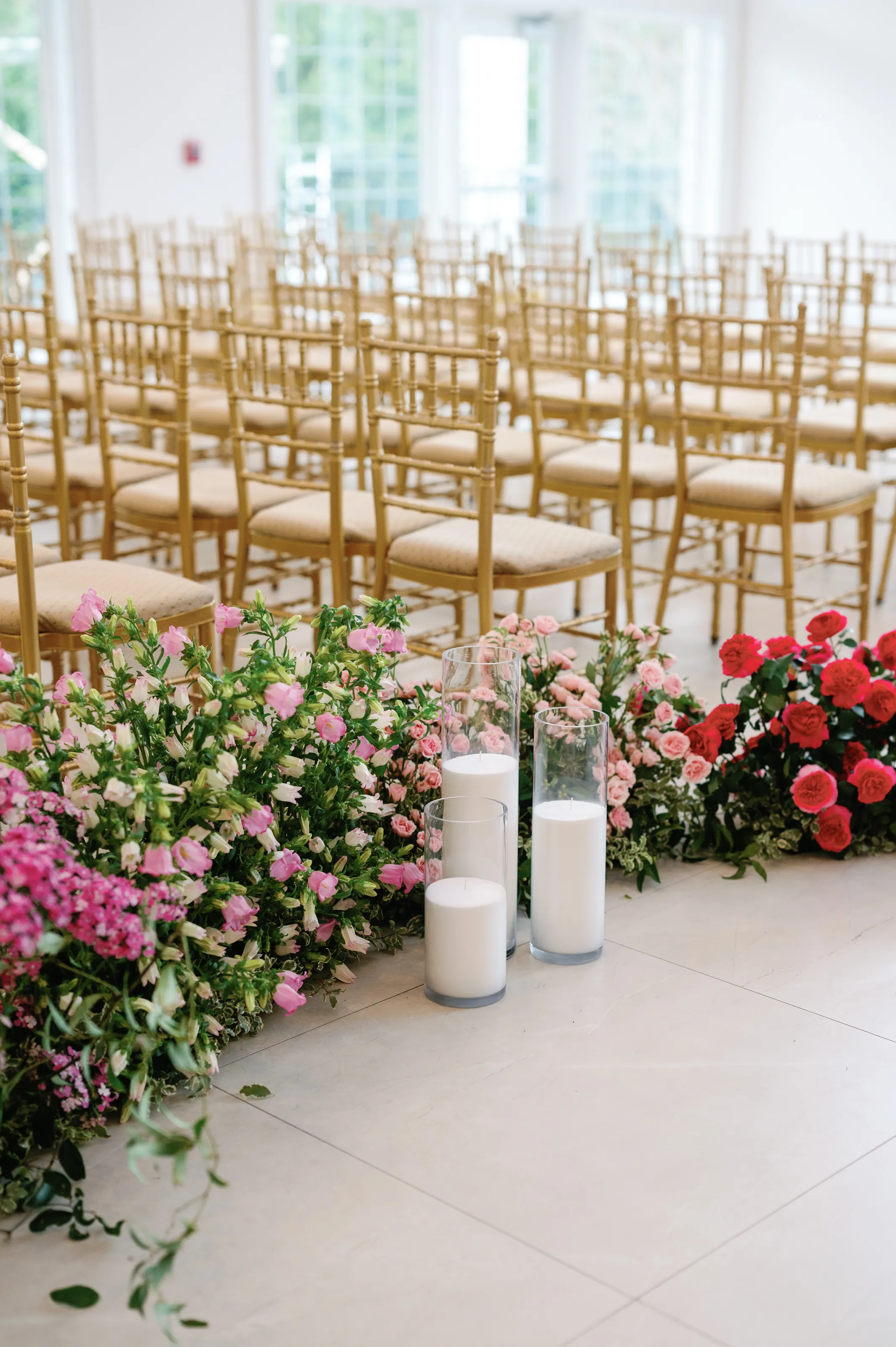 Rows of gold chiavari chairs frame a ceremony aisle lined with cascading pink and red rose runners and pillar candles at Estate at Florentine Gardens
