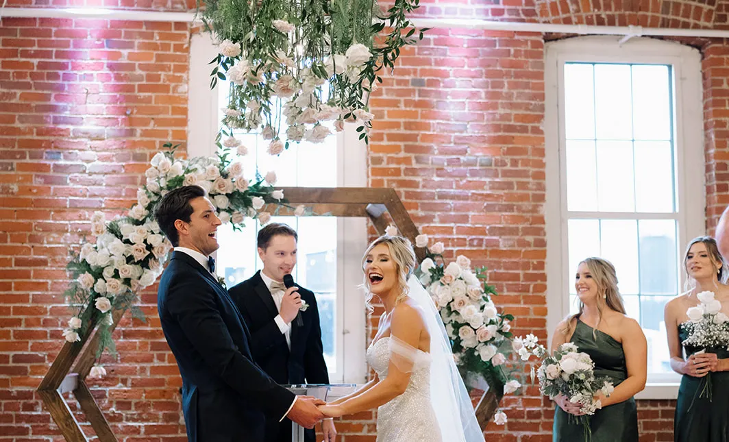 Bride and groom laughing together during their ceremony beneath a floral installation, exposed brick behind them