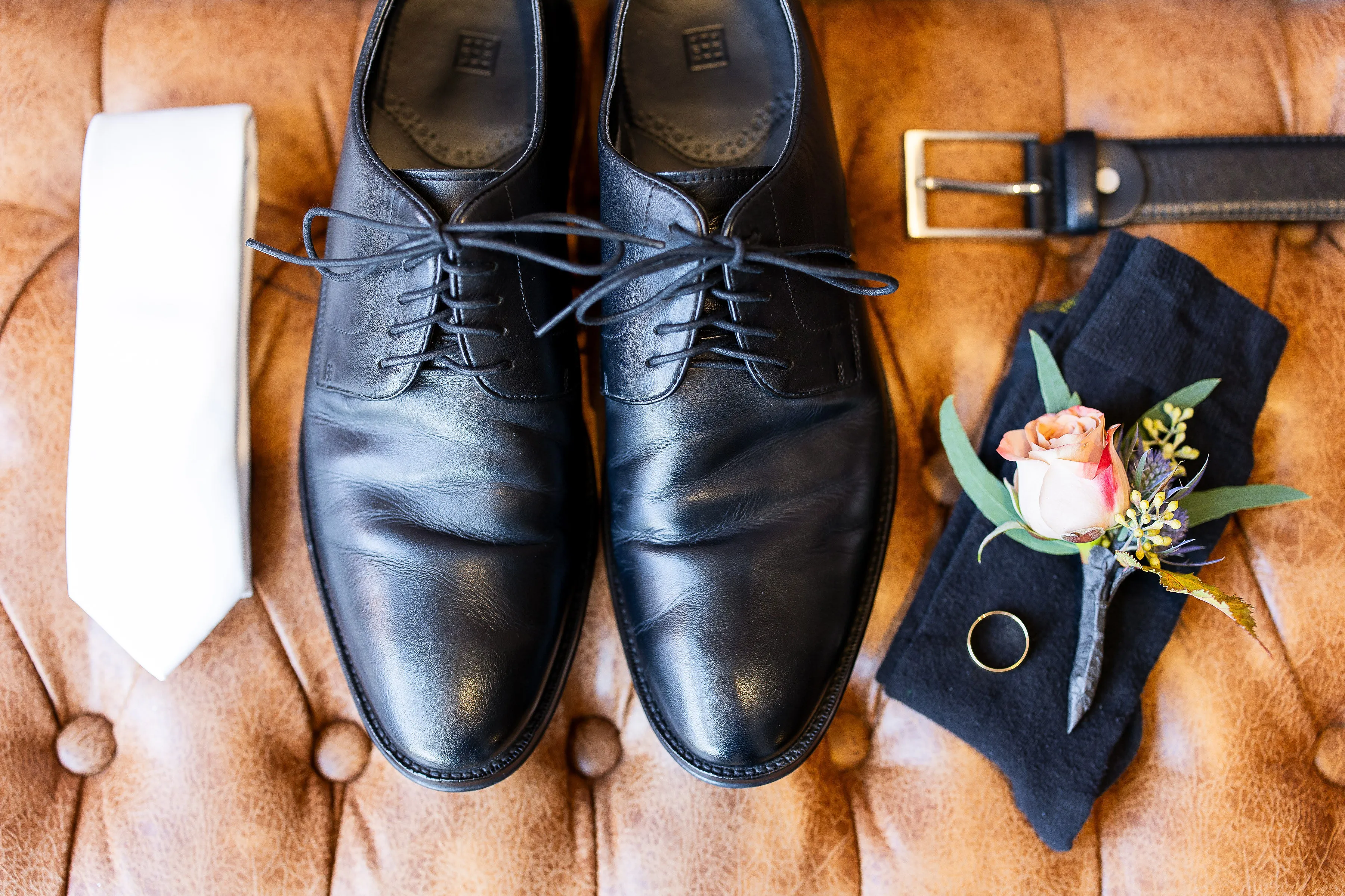 Nick's wedding day details — black shoes, tie, belt, and a blush rose boutonniere laid out before the ceremony