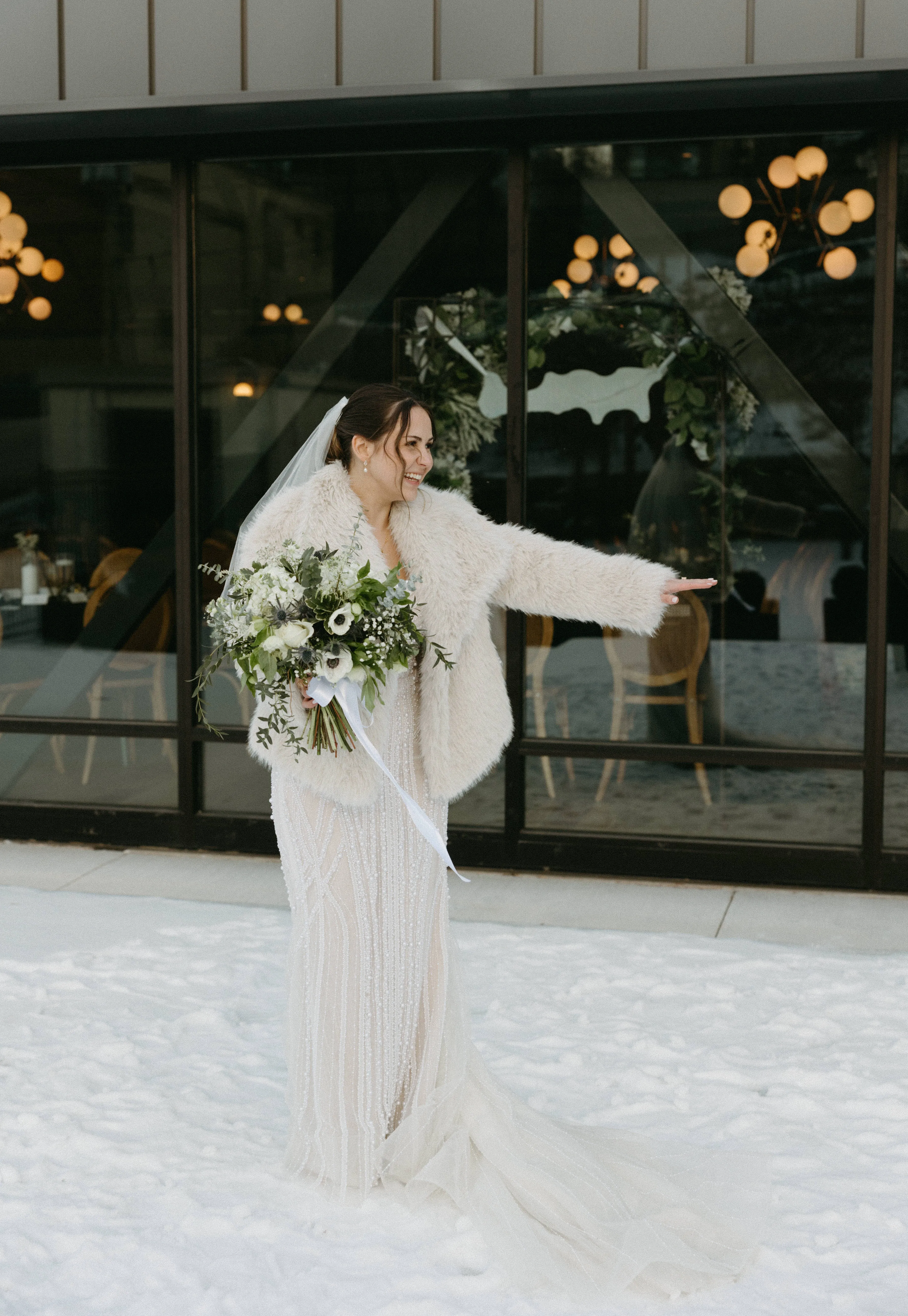Allie laughing in her fur coat against The Lora's glass facade, snow underfoot