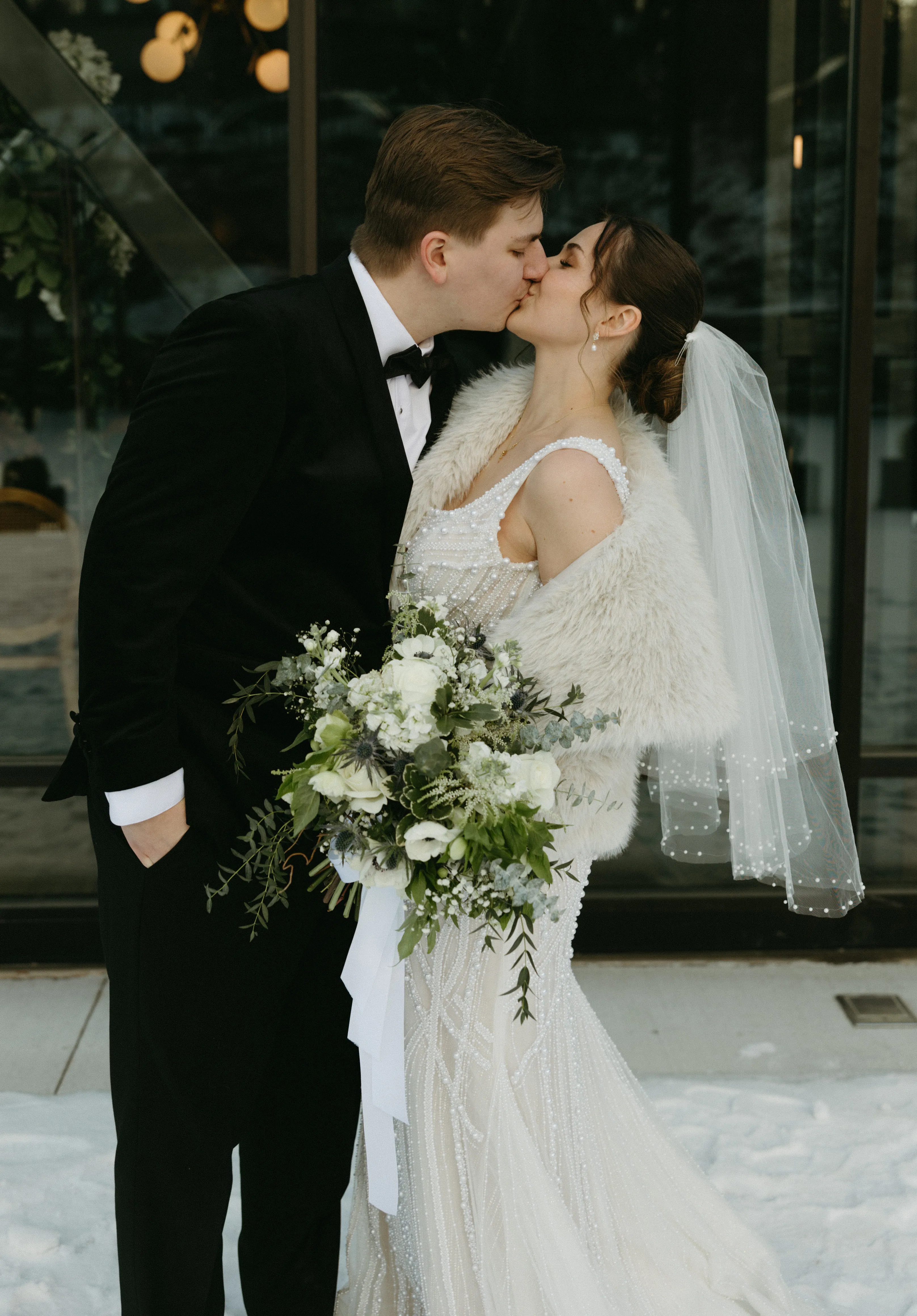 Allie and Joey kiss, her fur stole and veil catching the winter light, bouquet of white blooms and thistle between them