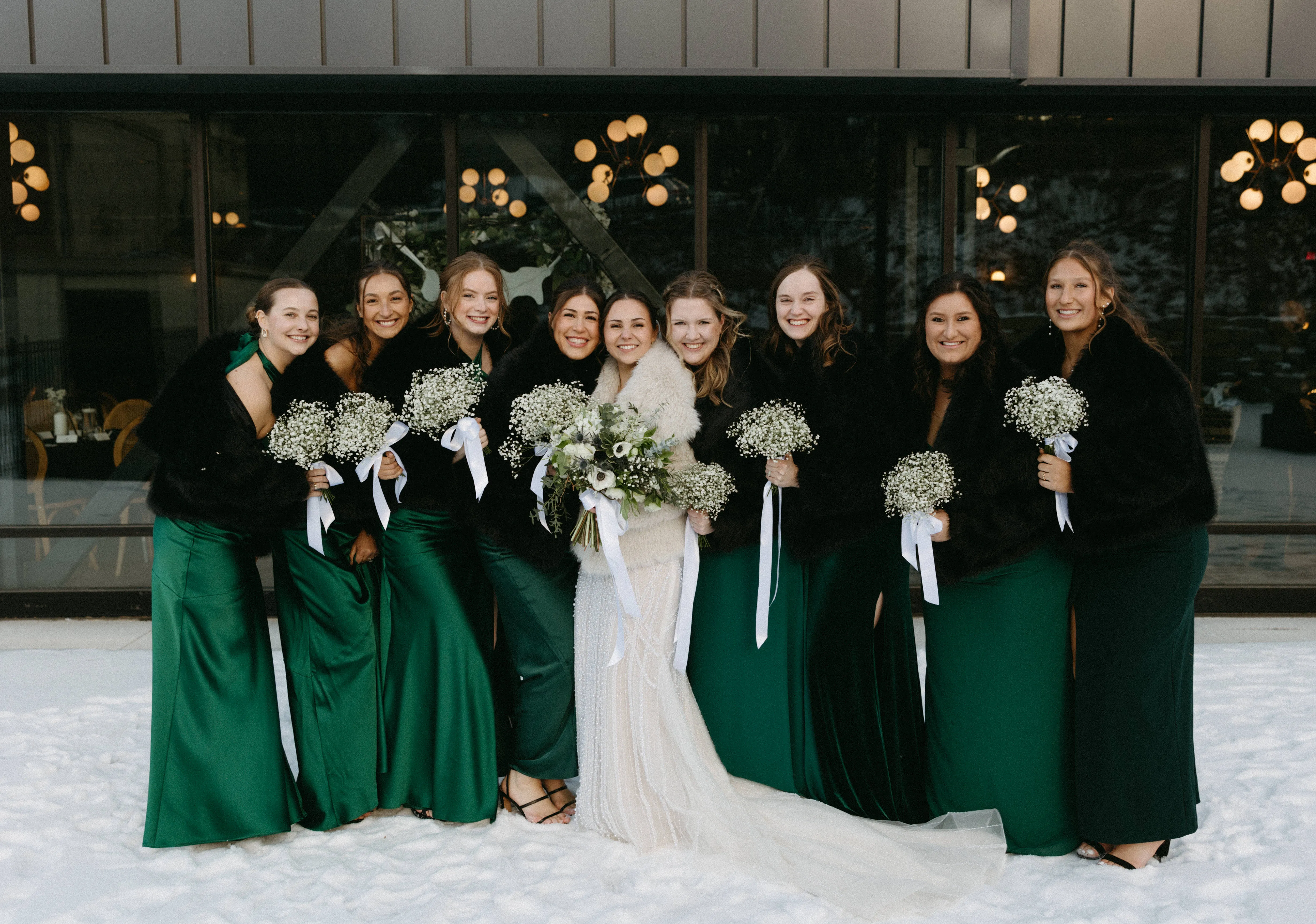 Allie and Joey's wedding party gathered in the snow outside The Lora, bridesmaids in emerald with fur stoles, bouquets of baby's breath