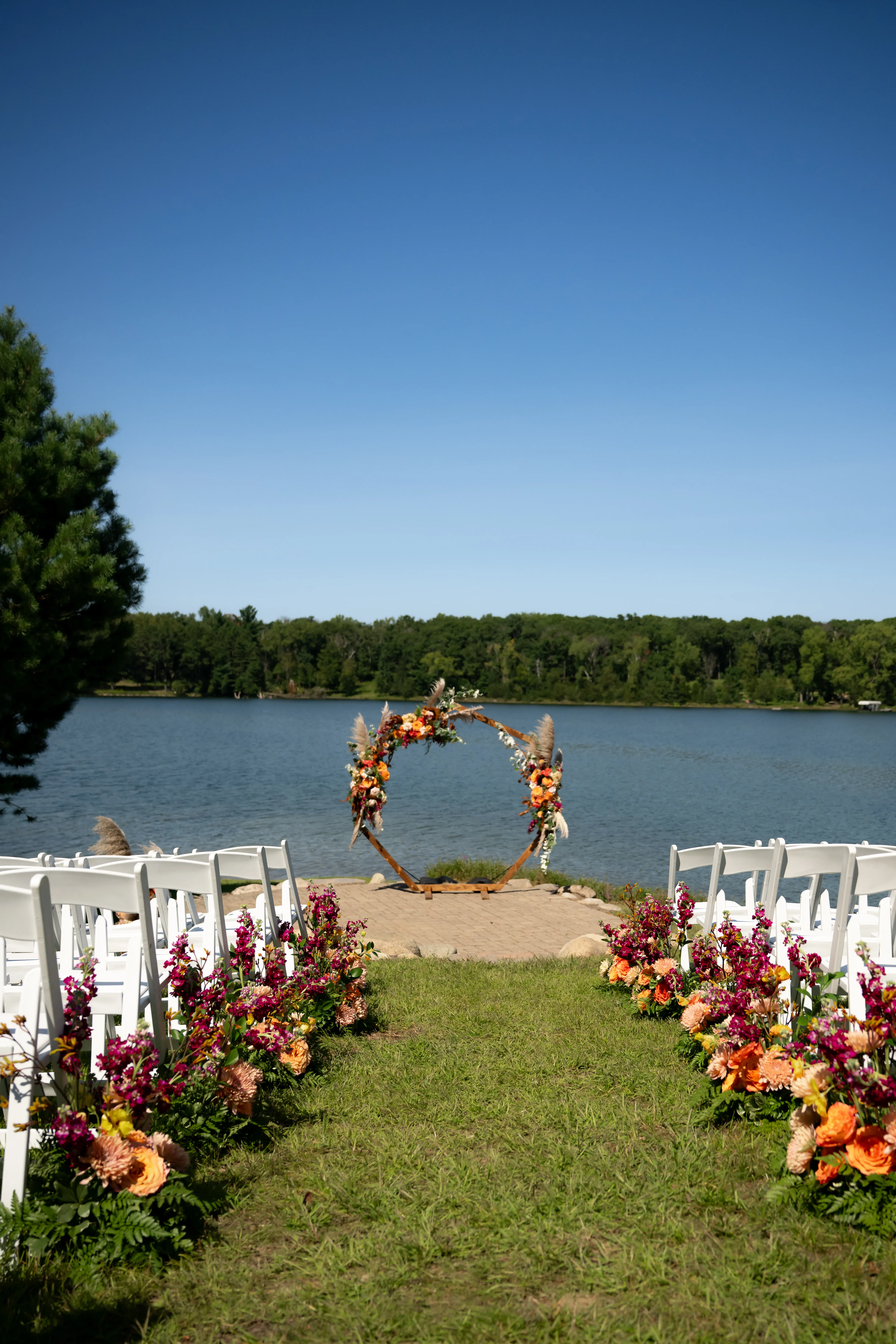 Wide view of the lakefront ceremony aisle, a hexagon arbor in a bold late-summer palette overlooking the water